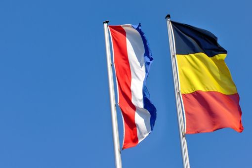 A flag of Belgium and one of the Netherlands under a blue sky.